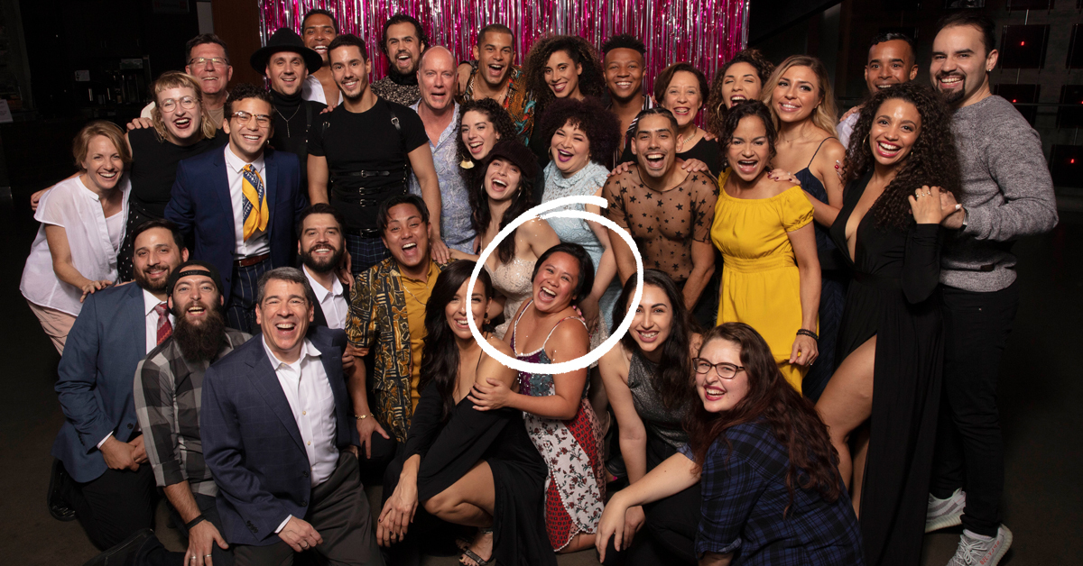 Kristen Mun front and center (like any good stage manager!) wearing a red and white dress and a huge smile. She’s surrounded by other members of the cast and creative team of In the Heights at the opening night reception.