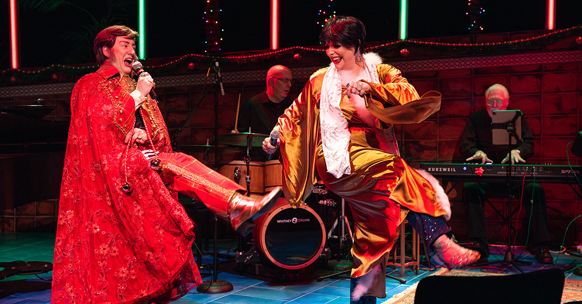 A man in a red cape and a smiling woman in a gold dress sing and kick their feet in unison while holding microphones.