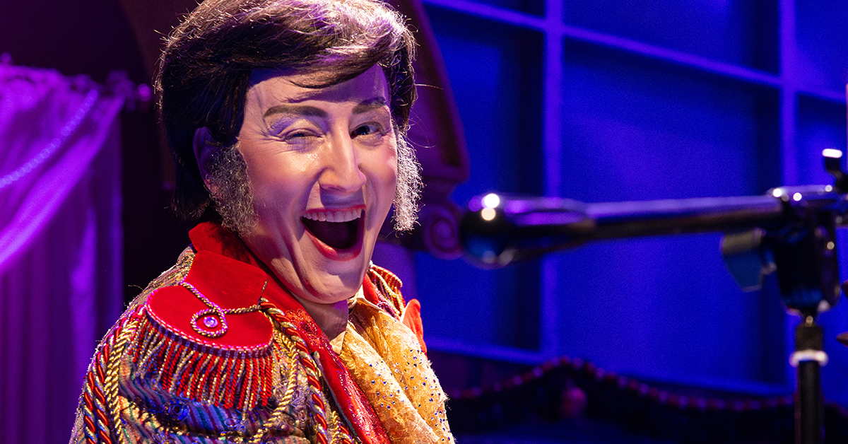 A man with mutton chop sideburns and a red & gold coat is seated at a piano and singing joyfully into a microphone.