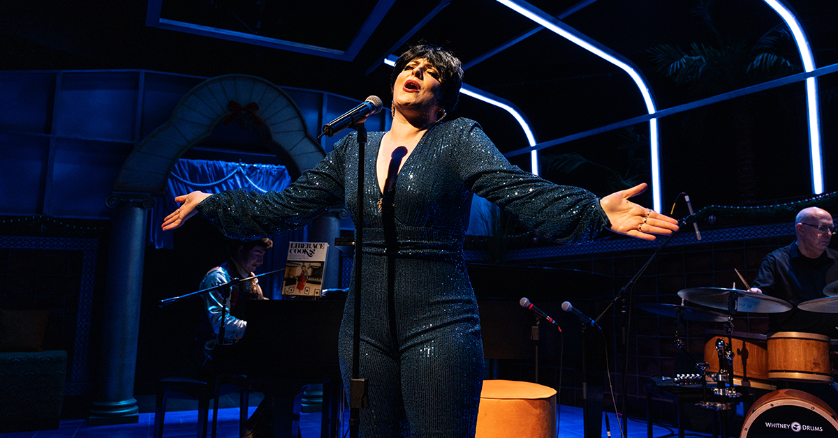 A woman in a dark blue sequined body suit stands singing at a microphone, her arms stretched wide.