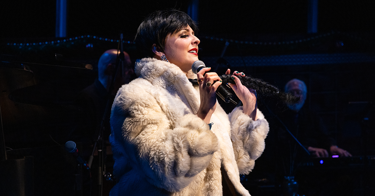 A woman wrapped in a fur coat sings into a mic, smiling with closed eyes and holding a long braid of her hair.