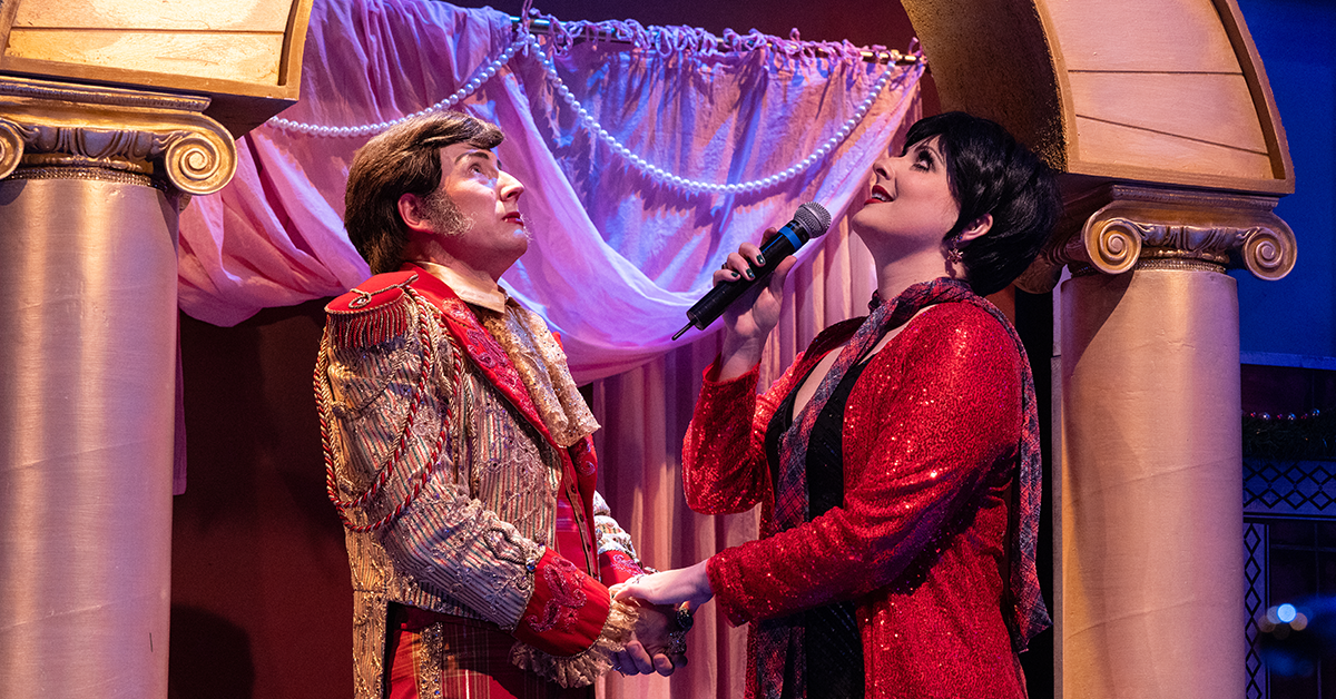 A man in a red & gold coat and a singing woman in a red sequined top hold hands and look up in front of a decorative archway.