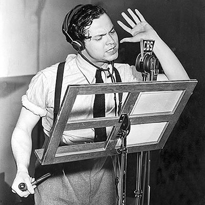 A man wearing headphones stands at a radio microphone, reading aloud from a script on a music stand, one hand raised dramatically.