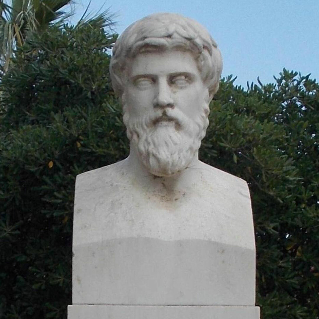 A white marble bust of a bearded man in front of green foliage and a blue sky.