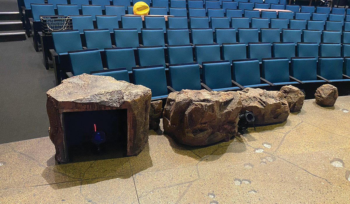A set viewed from the POV of someone onstage reveals hollow false rocks that conceal lighting equipment. Rows of empty seats lie beyond.
