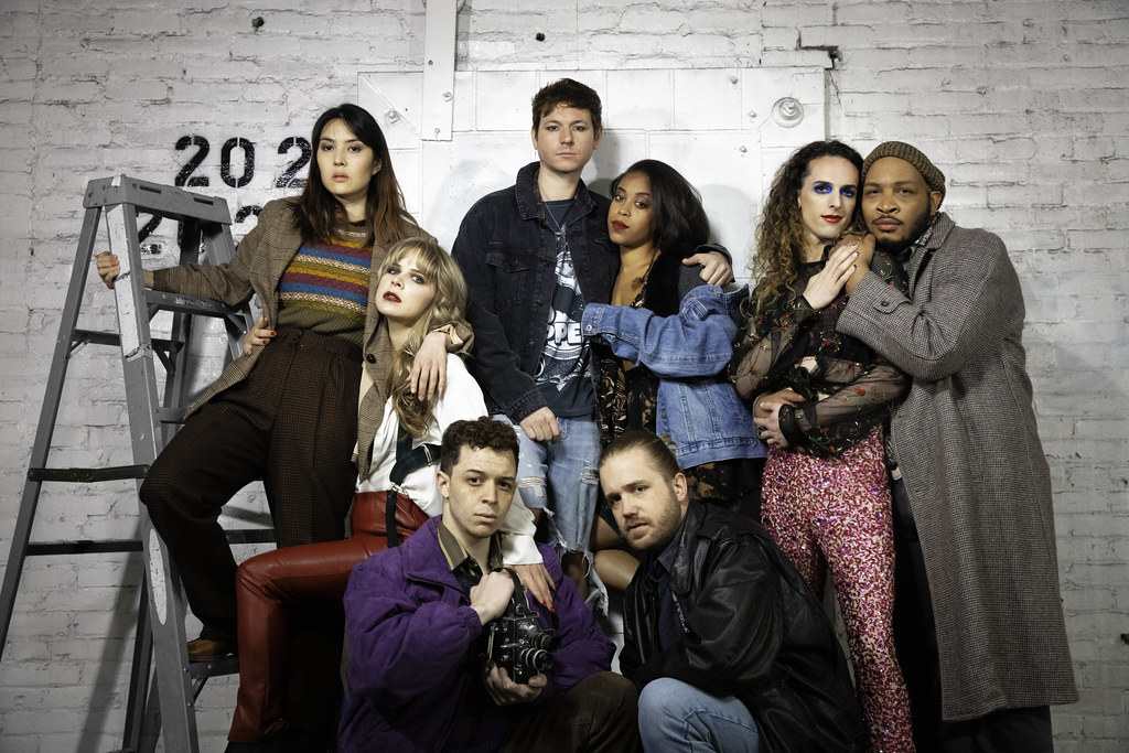 A group of eight people stand in front of a white brick wall; they are huddled together, staring into the camera.