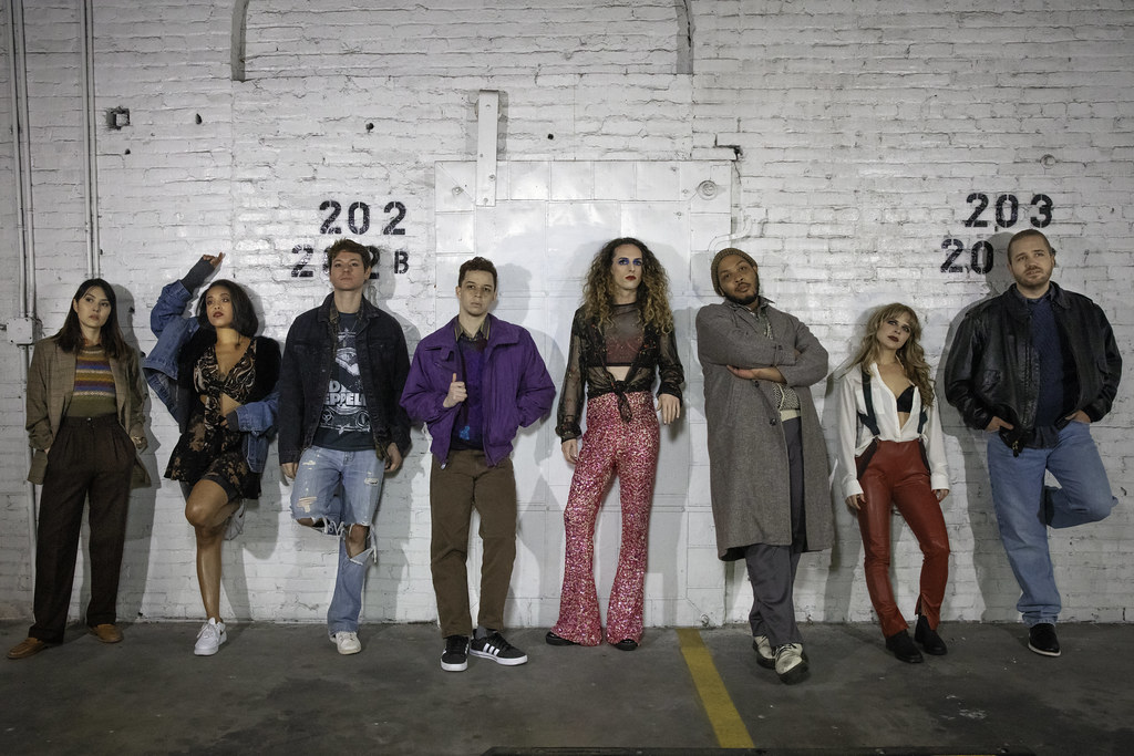 A group of eight people lean against a white brick wall; they look intently off into the middle distance.