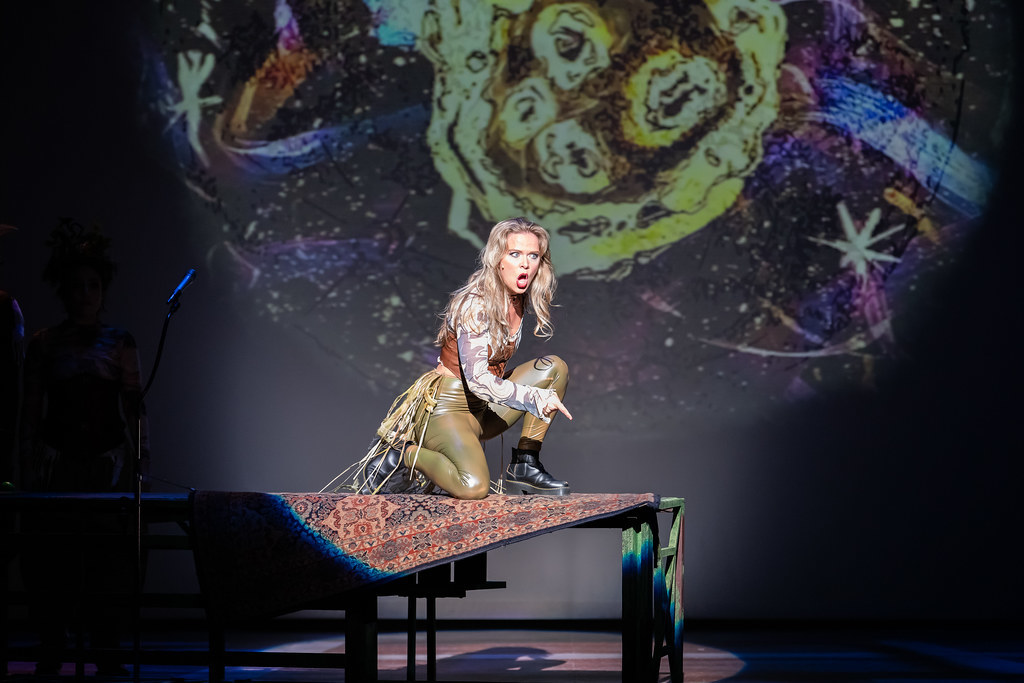 A woman kneels on a small stage platform pointing down, passionately mid-speech. 