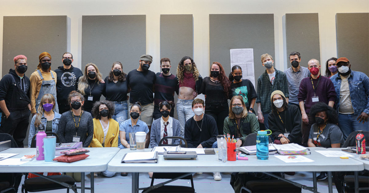 A group of twenty-four people in masks smile at the camera from behind a a long table.