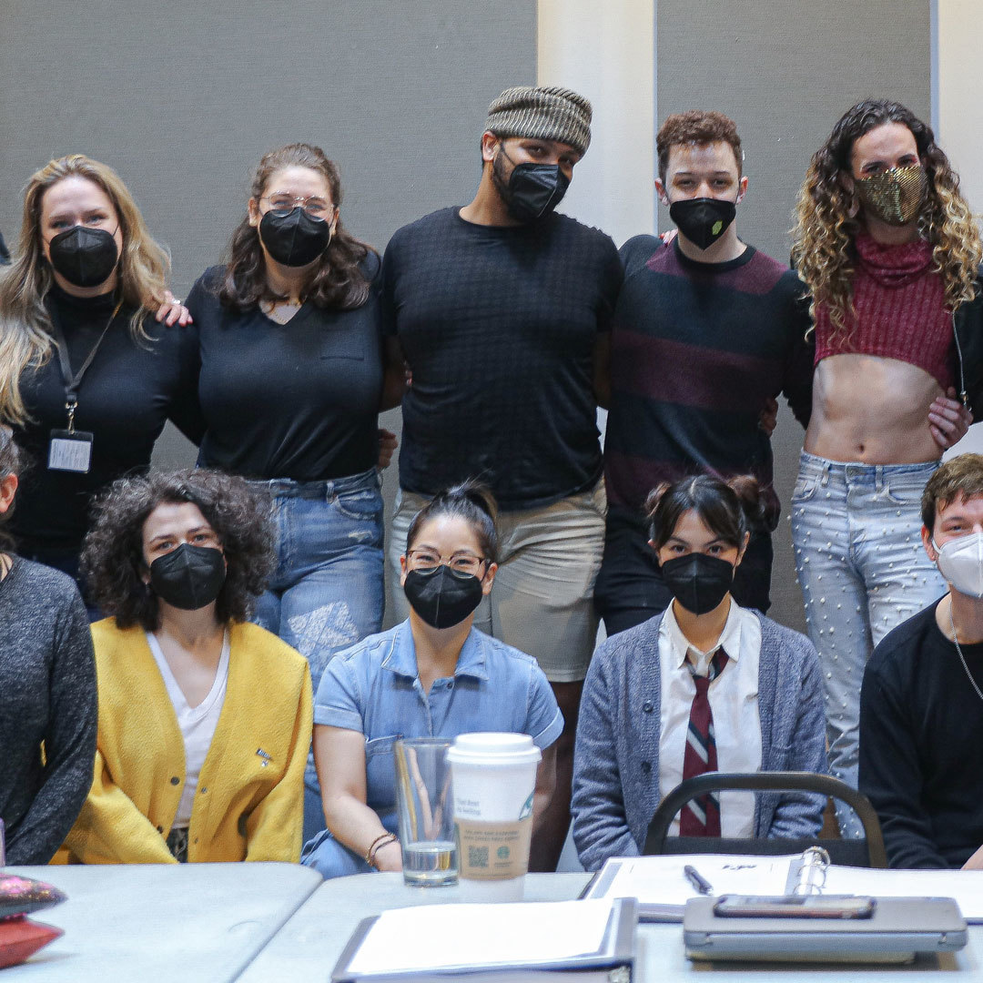 A group of twenty-four people in masks smile at the camera from behind a a long table.