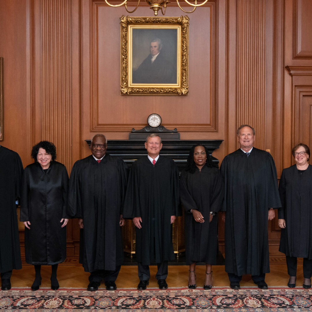 Smiling men and women wearing black robes pose side-by-side in a wood-paneled room; image is cropped so only 6 of 9 justices are visible.