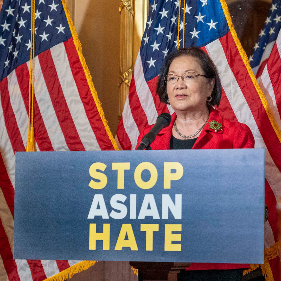 A woman speaks in front of a row of American flags behind a podium that prominently displays the words "Stop Asian Hate."