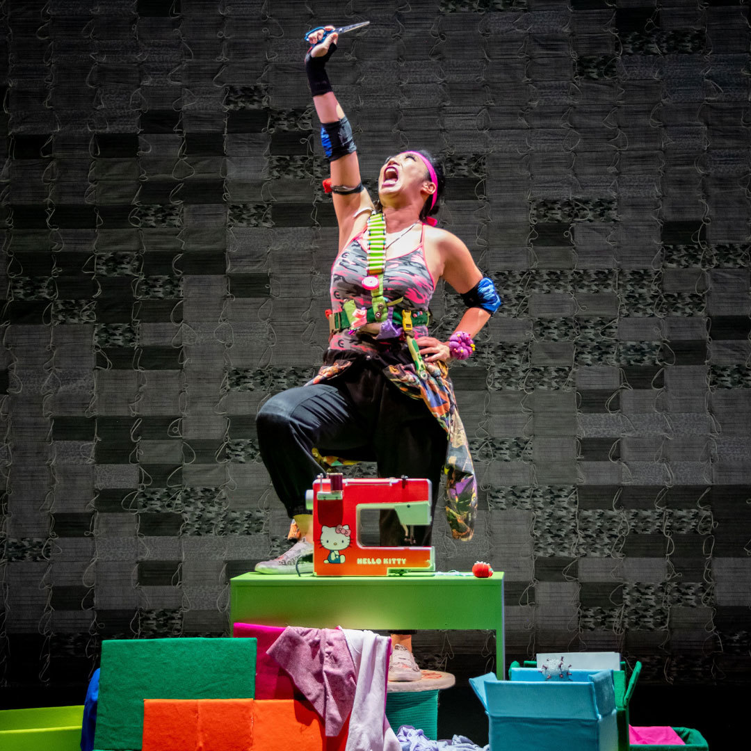 A woman stands triumphantly on a pile of boxes and sewing instruments; she is holding aloft a pair of scissors.