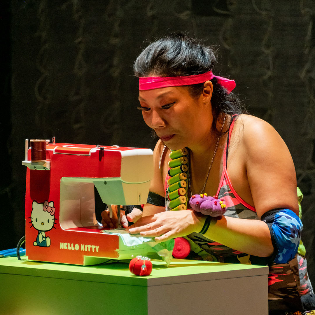 A woman works diligently at a sewing machine making a mask; the sewing machine is branded with 'Hello Kitty.'