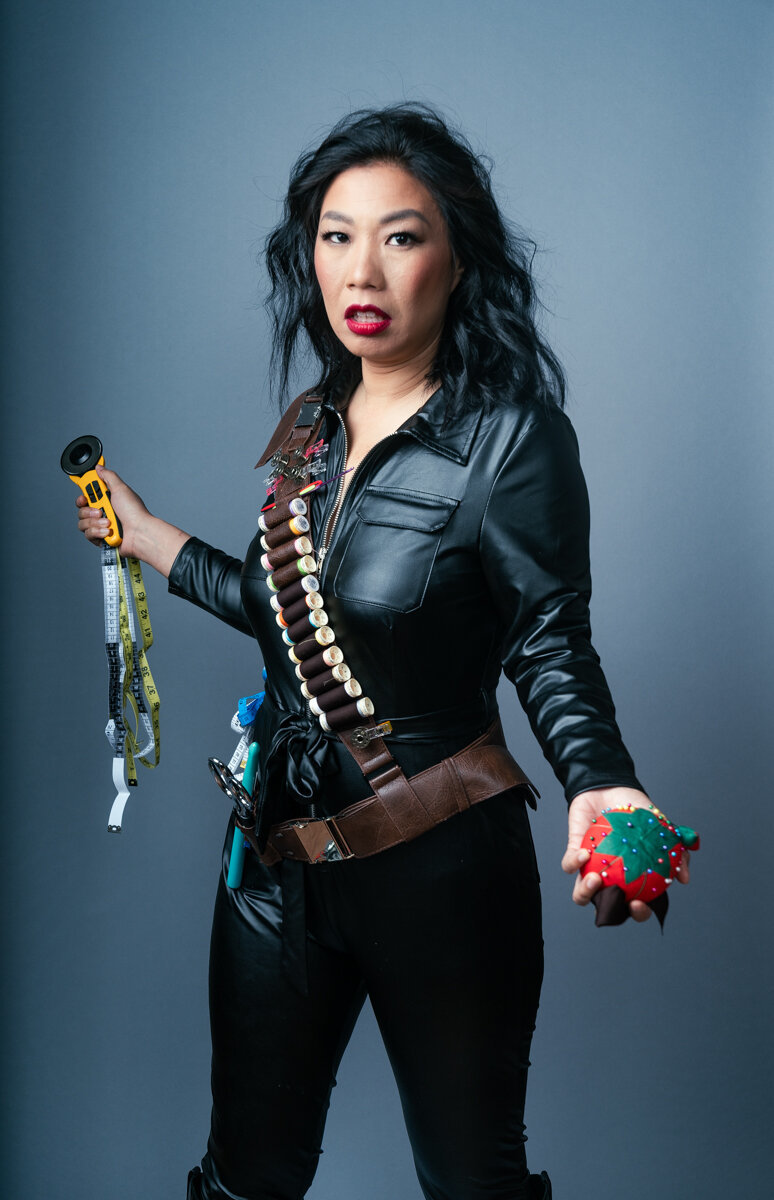 A woman wearing a leather jumpsuit stares at the camera brandishing a tomato pin cushion and sewing tool 