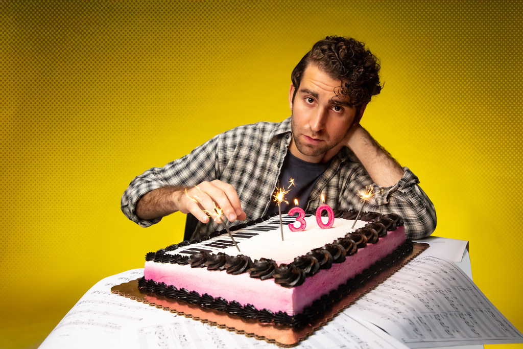 A man hesitantly hovers above the top of. cake designed like a piano; sparklers are glowing brightly on top of the cake.