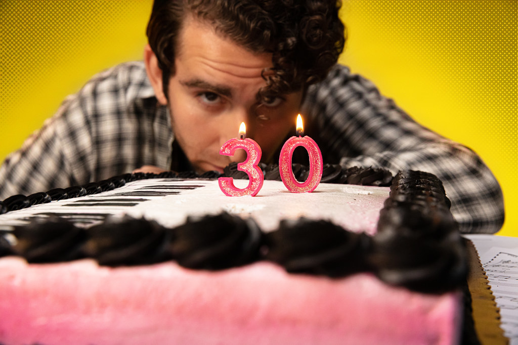 A man stares deeply into the flames on top of two candles in a cake; the candles read "3, 0".
