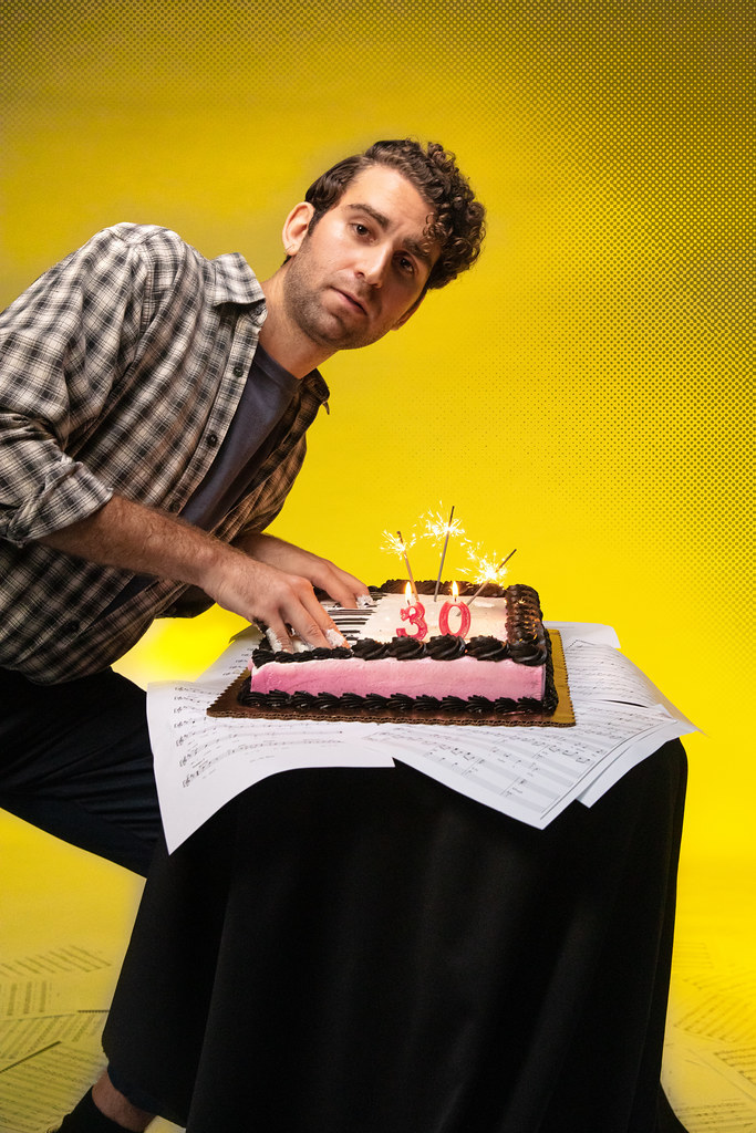 A man sticks his fingers into a cake as tho he is playing a piano; he looks banal and nonplused.