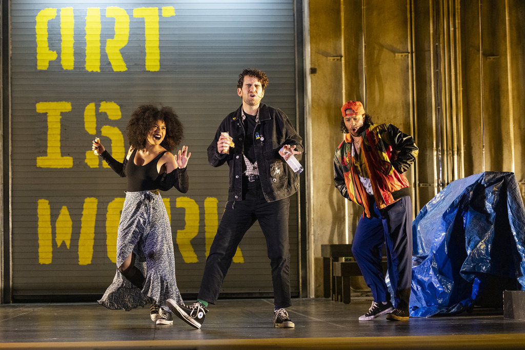 Three people in cool 90s clothing dancing and singing in front of a wall that reads ART IS WORK.