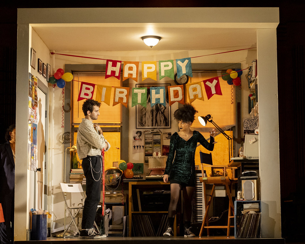 Two people in cool 90s clothing looking at each other across a room and are standing under flags that read Happy Birthday.