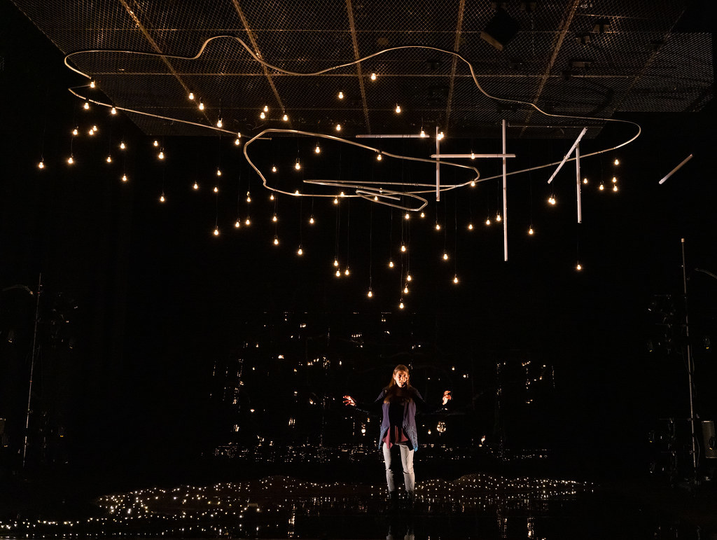 A woman stands amongst a dazzling array of fairy lights that engulf the stage around her.