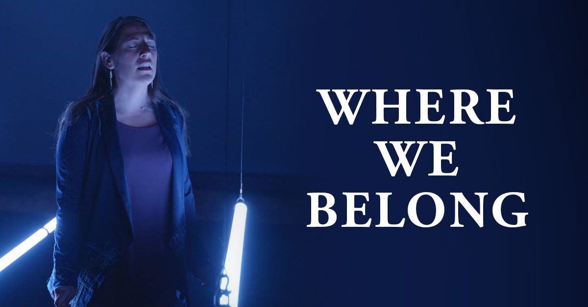 The title "Where We Belong" overlaid on a photo of a woman, her eyes closed, standing between fluorescent lights hanging at waist level.