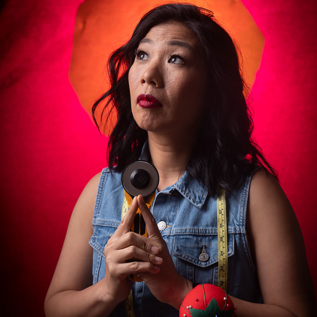 A woman with dark hair holding sewing implements in front of a dramatic red-orange background, gazing pensively up and to the right.