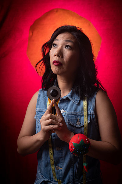 A woman with dark hair holding sewing implements in front of a dramatic red-orange background, gazing pensively up and to the right.