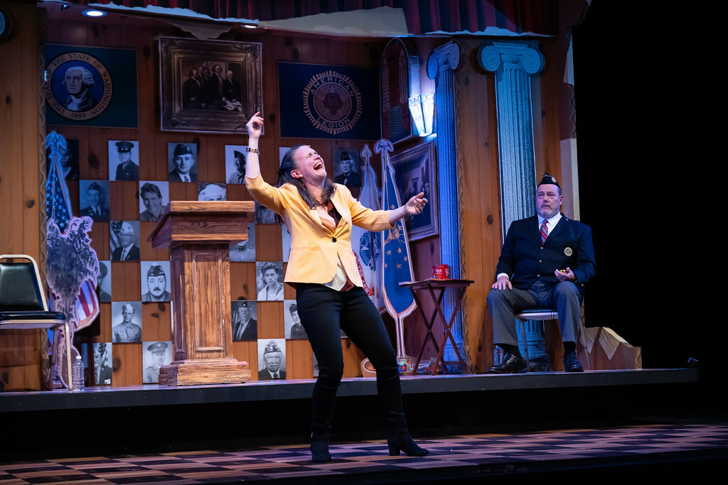 A woman stands, her head thrown back, gesturing wildly as seated man in a legionnaire's uniform looks on.