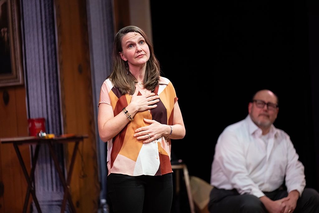 A woman stands gesturing with her arms crossing her chest as seated man looks on.