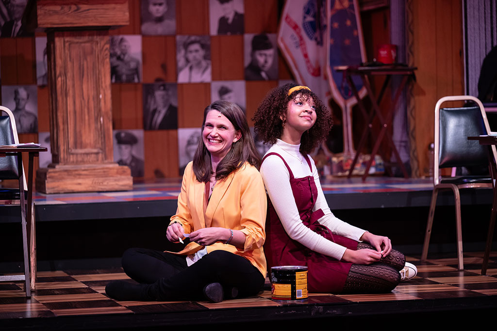 A woman and a young woman sit on the floor with their backs against each other.