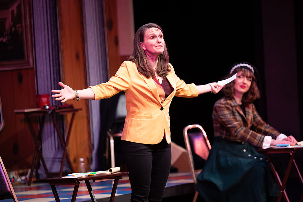 A woman stands gesturing with her arms wide as a seated younger person looks on.