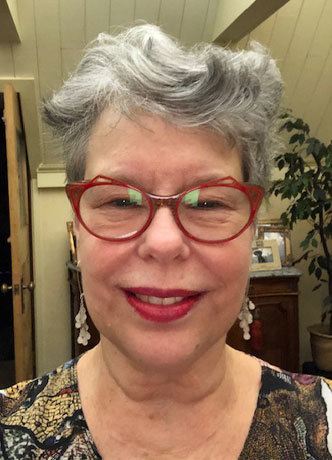 White woman smiling with red glasses and grey short cut hair. 