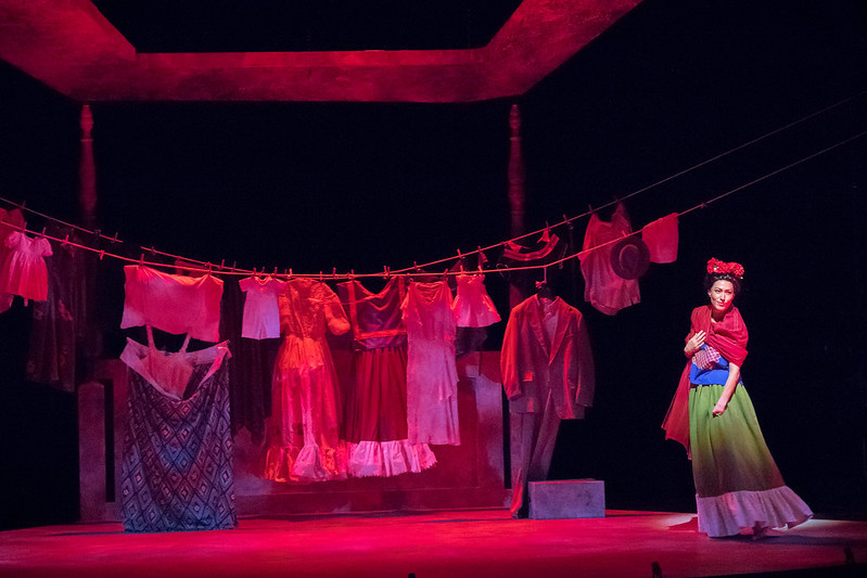 A variety of outfits are strung up on a clothing illuminated by a red wash of light. Vanessa Severo as Frida Kahlo stands on 