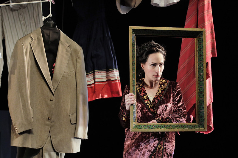 Vanessa Severo as Frida beside a tan suit hanging, wearing a maroon crushed velvet robe and holding a gold frame which she is
