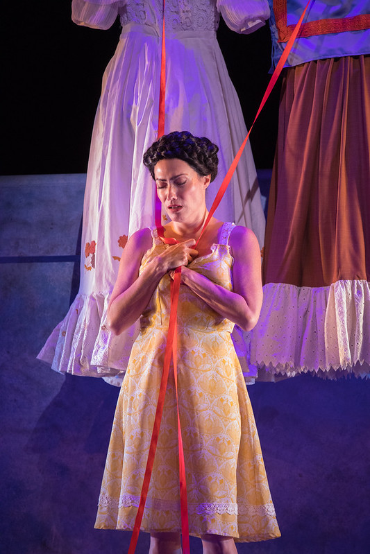 Vanessa Severo as Frida, wearing a yellow and white sun dress. On a clothesline to her left hangs a white dress, on the right