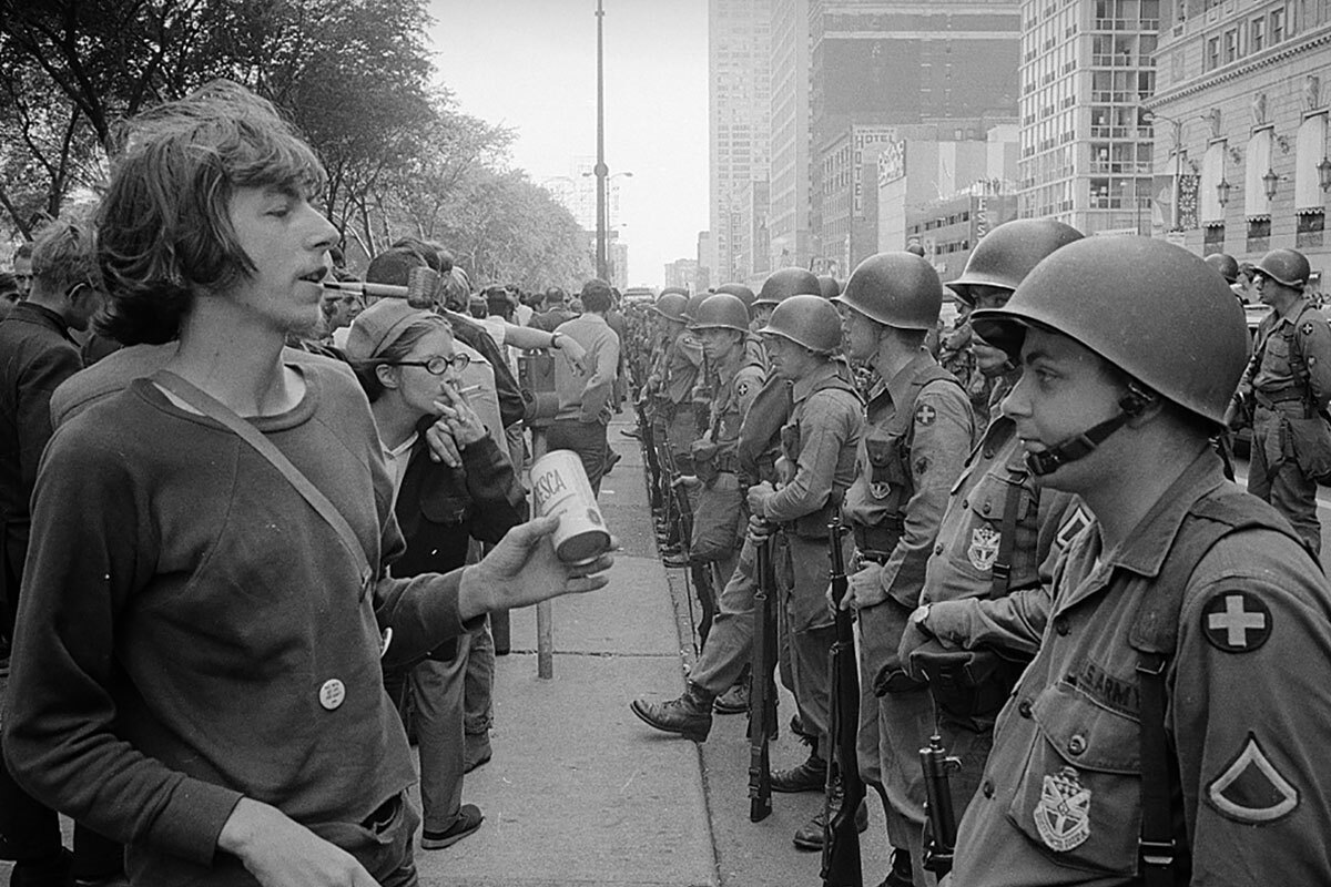 A gathering of young, long-haired war protesters on a city sidewalk are bordered by a row of armed soldiers along the adjacent street.