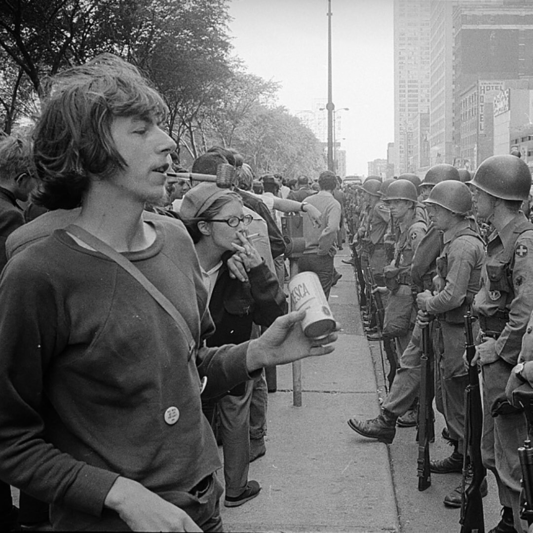 A gathering of young, long-haired war protesters on a city sidewalk are bordered by a row of armed soldiers along the adjacent street.