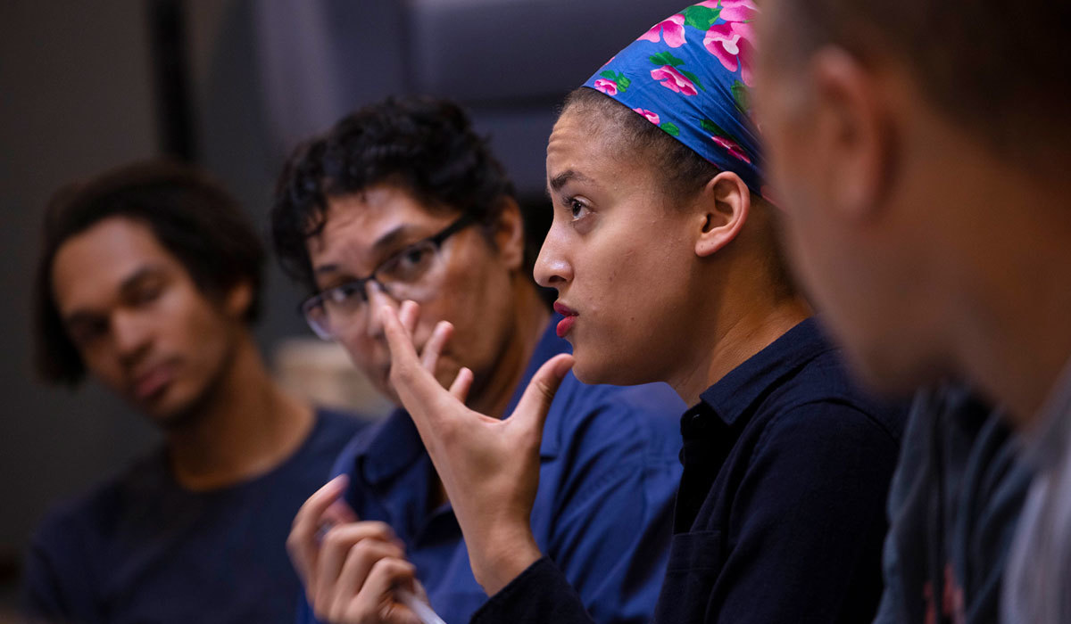 A person of color is speaking and gesturing with their hands as three other people look on.