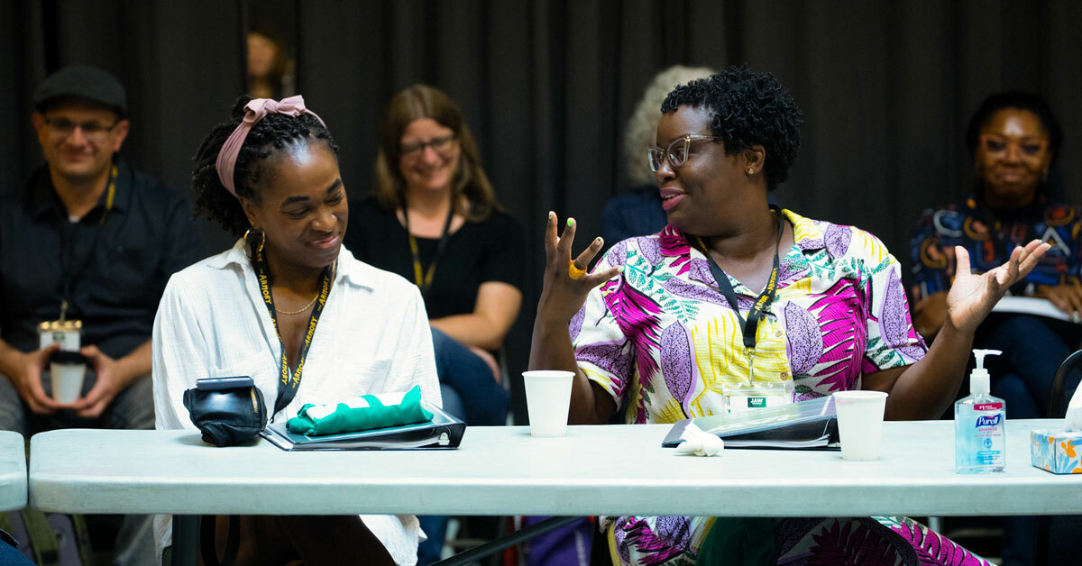 Two women sit at a work table gesturing and smiling as others look on in the background.