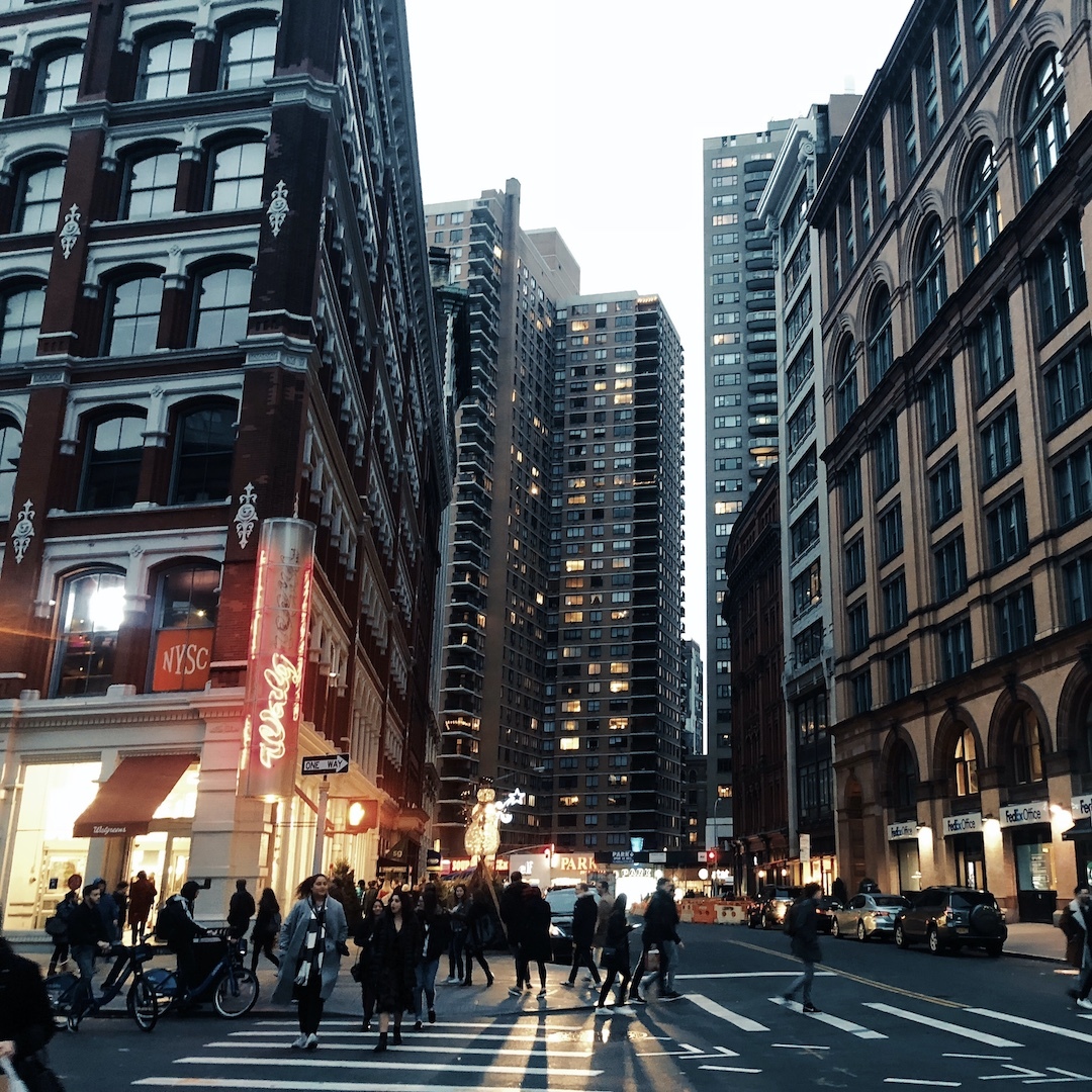 A busy Manhattan street in the evening, framed by lit shop windows and high-rise apartment buildings.