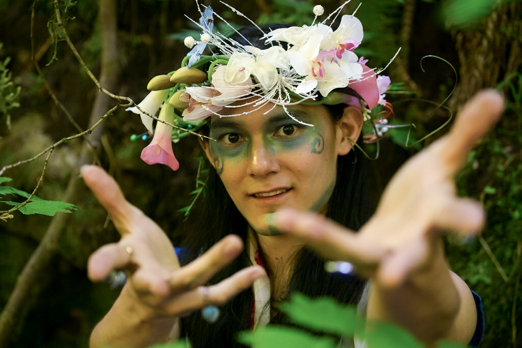 A man in a forest, with painted designs on his face and a wreath of flowers on his head gestures magically toward us.