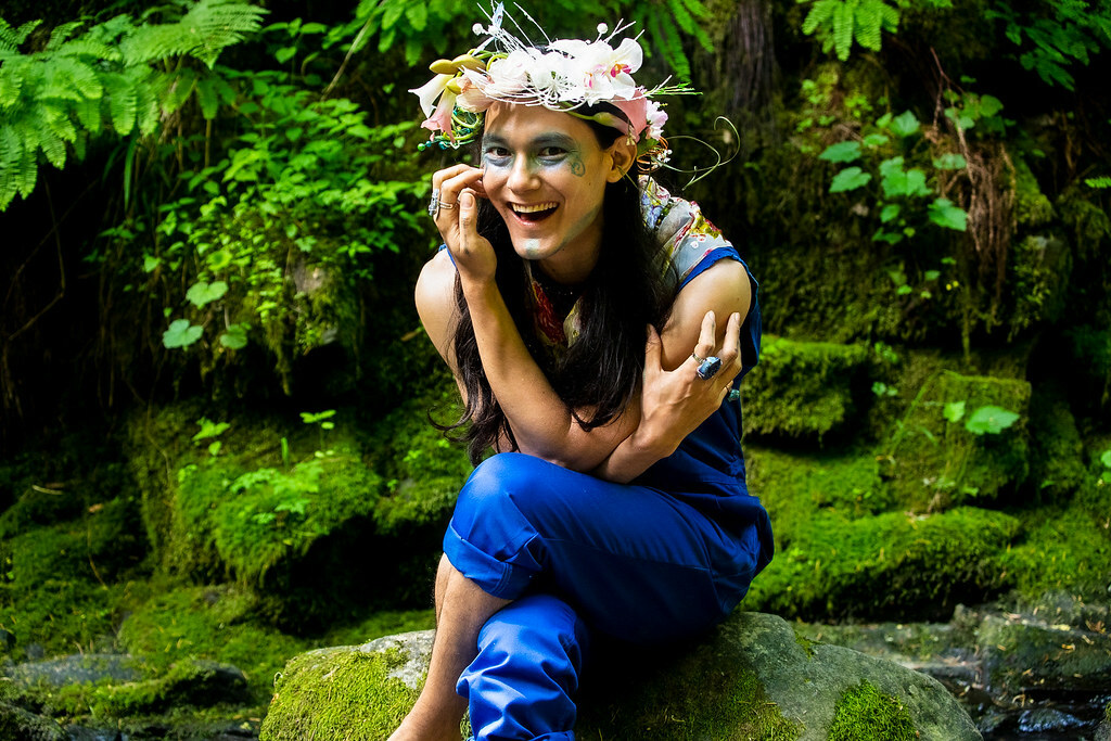 A man in a forest, wearing painted designs on his face and a wreath of flowers on his head sits on a mossy rock, smiling.