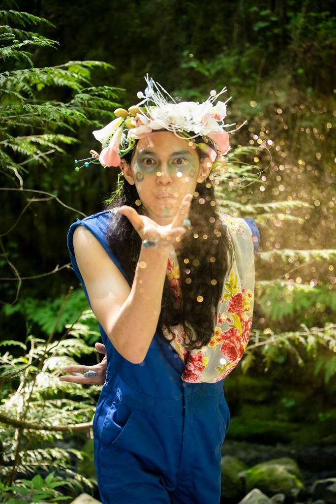 A man in a forest, wearing a wreath of flowers on his head, releases a cloud of glitter into the air.