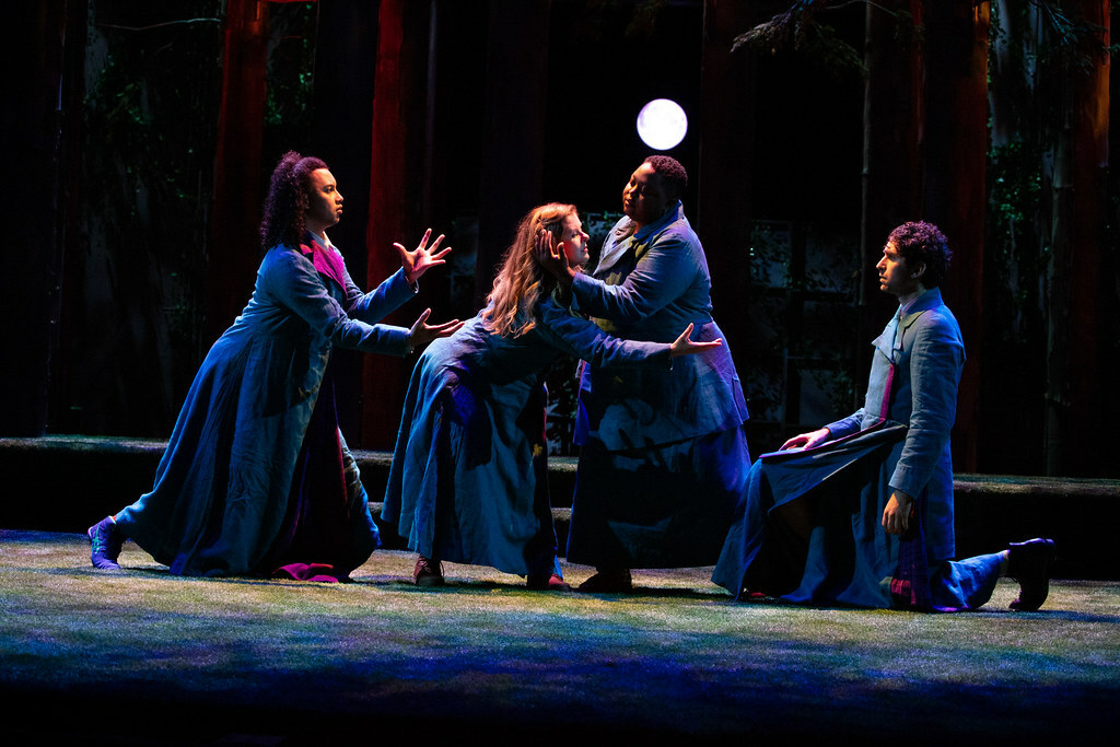 Four characters interact with each other in various stances on a dimly lit stage; trees and a full moon in the background.