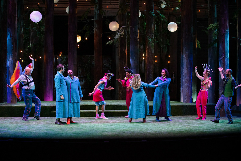 A group of nine colorfully dressed humans and fairies celebrate on the turf-covered stage beneath trees and twinkly lights. 