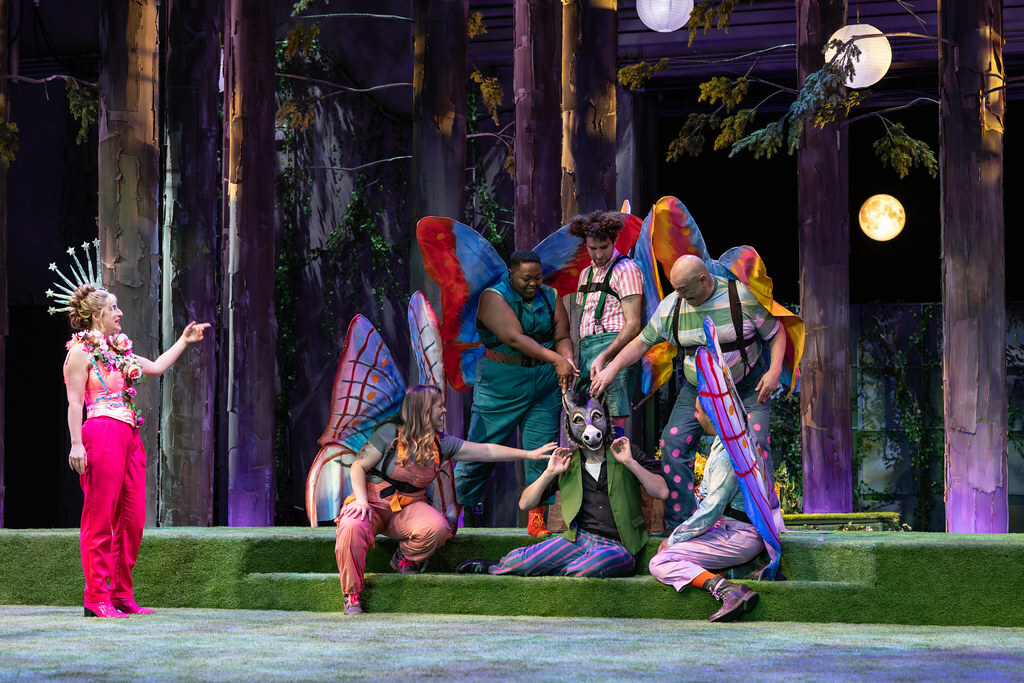 Among the trees on a turf-covered stage, a group of colorful fairies surround a man who has the head of a donkey.