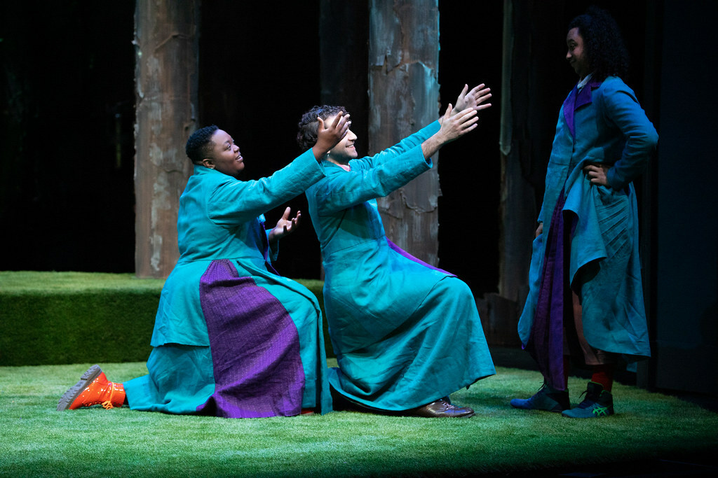 In front of some trees on a turf-covered stage, two people kneel and reach out their hands to a third person who is standing.