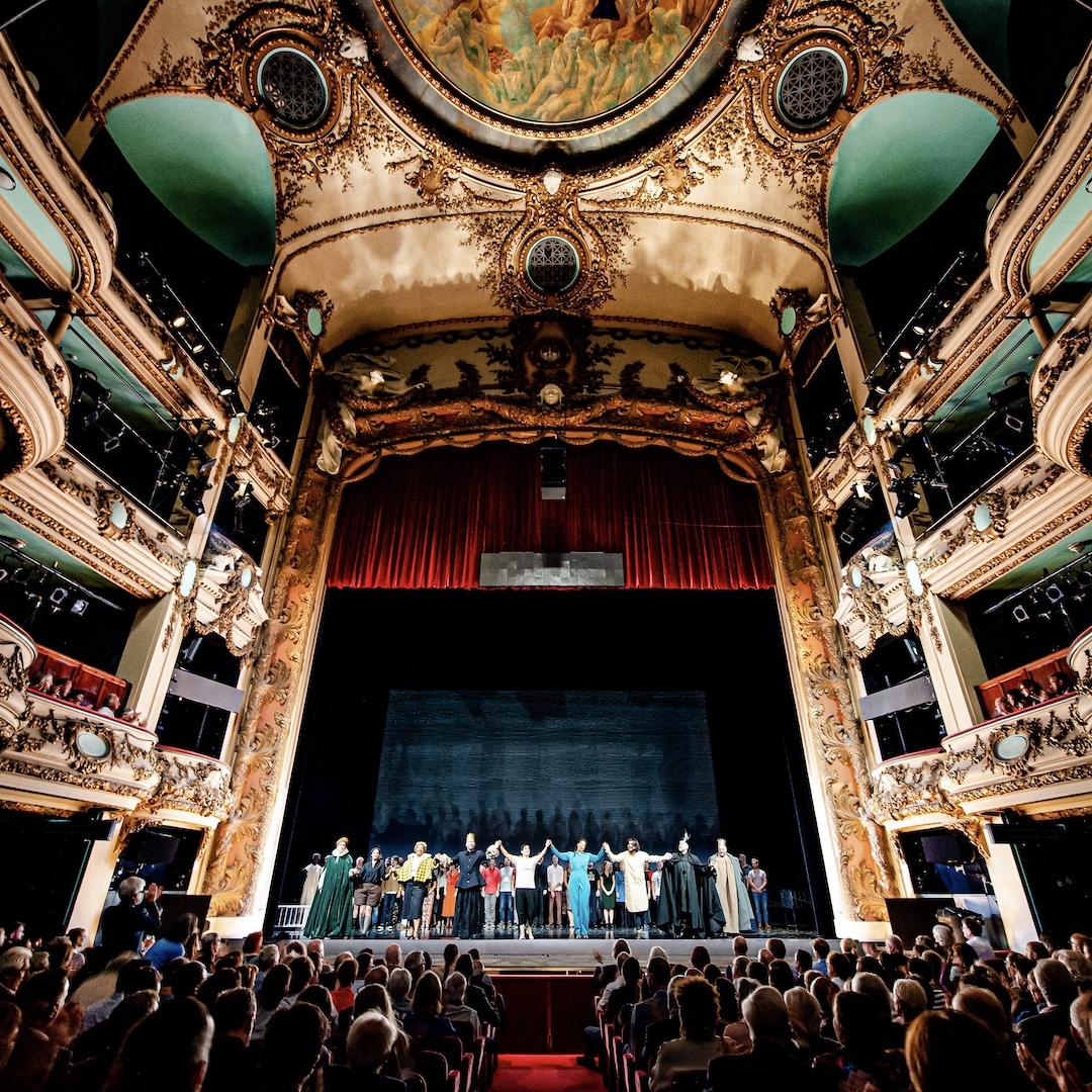 Floor-to-ceiling view of an ornate opera house with seated audience members in the foreground and performers on stage.