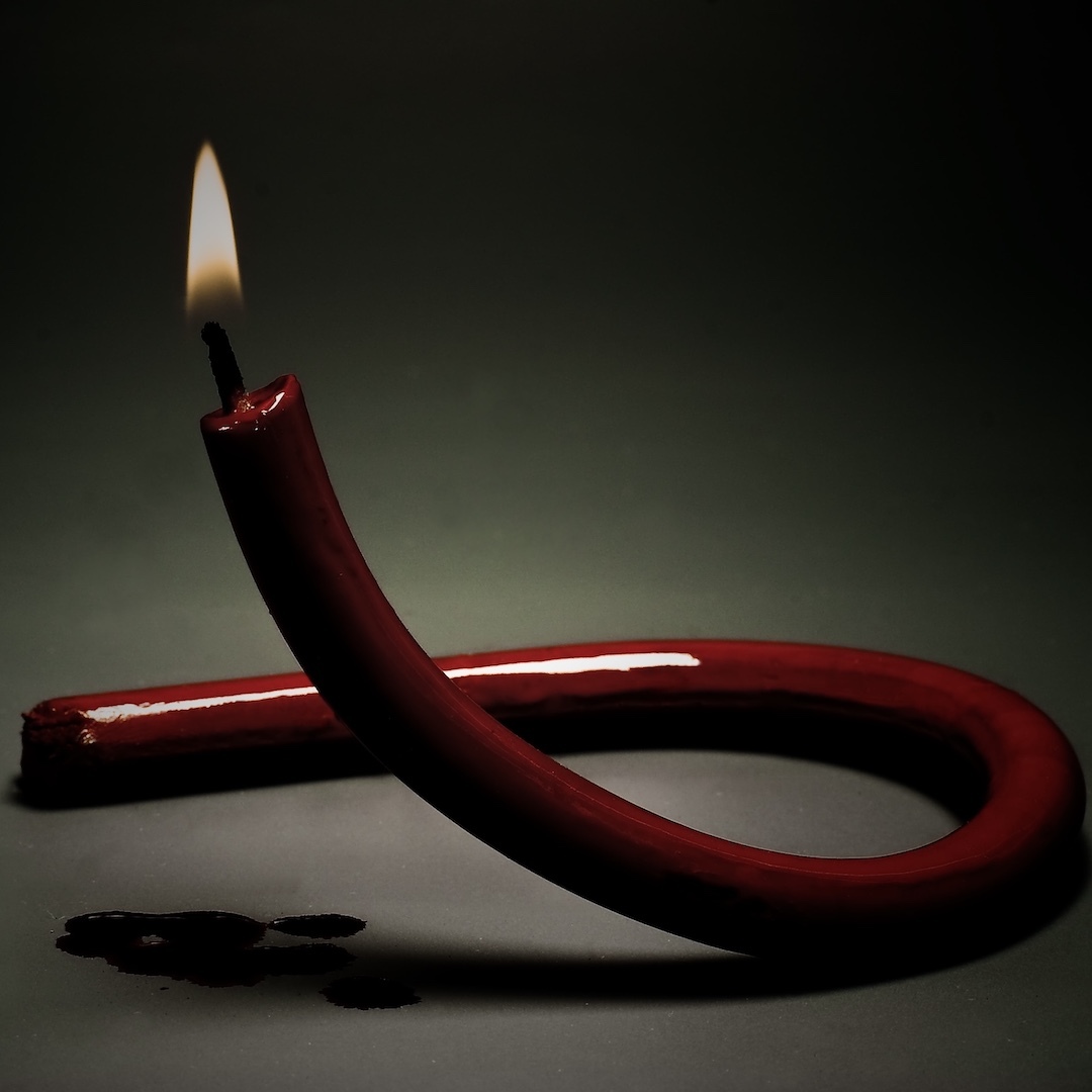 A lit, red wax candle, long and slender, that has been twisted into the shape of a red AIDS Awareness ribbon.